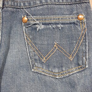 May include: Close-up of a blue denim jeans pocket. The pocket features a classic design with a stitched 'W' pattern in gold thread. Two copper-colored rivets secure the pocket to the jeans. The denim shows signs of wear.