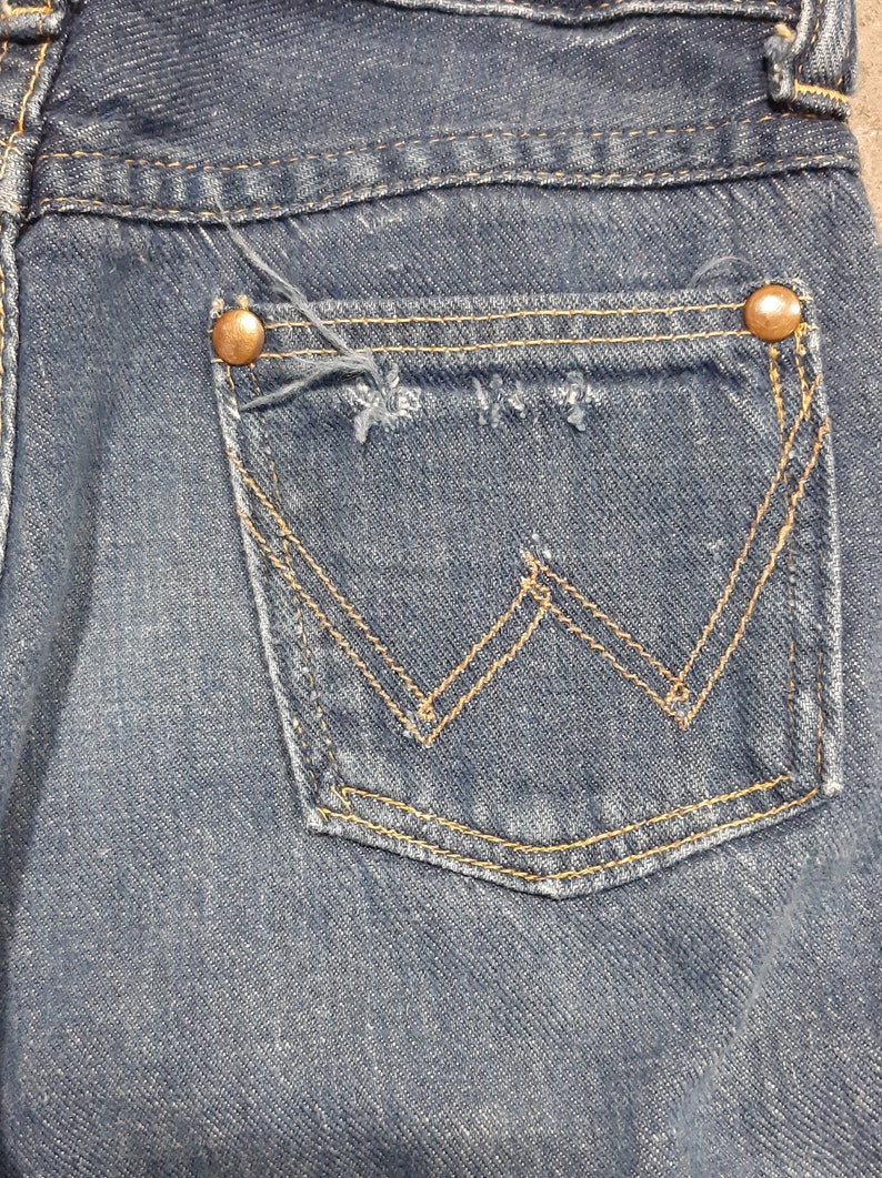 May include: Close-up of a blue denim jeans pocket. The pocket features a classic design with a stitched 'W' pattern in gold thread. Two copper-colored rivets secure the pocket to the jeans. The denim shows signs of wear.