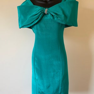 Puede incluir: Vestido de c&oacute;ctel color turquesa con escote de hombros descubiertos y un gran lazo en el busto. Un broche de pedrer&iacute;a brillante adorna el lazo. El vestido entallado, a la altura de la rodilla, se exhibe en un maniqu&iacute; negro.