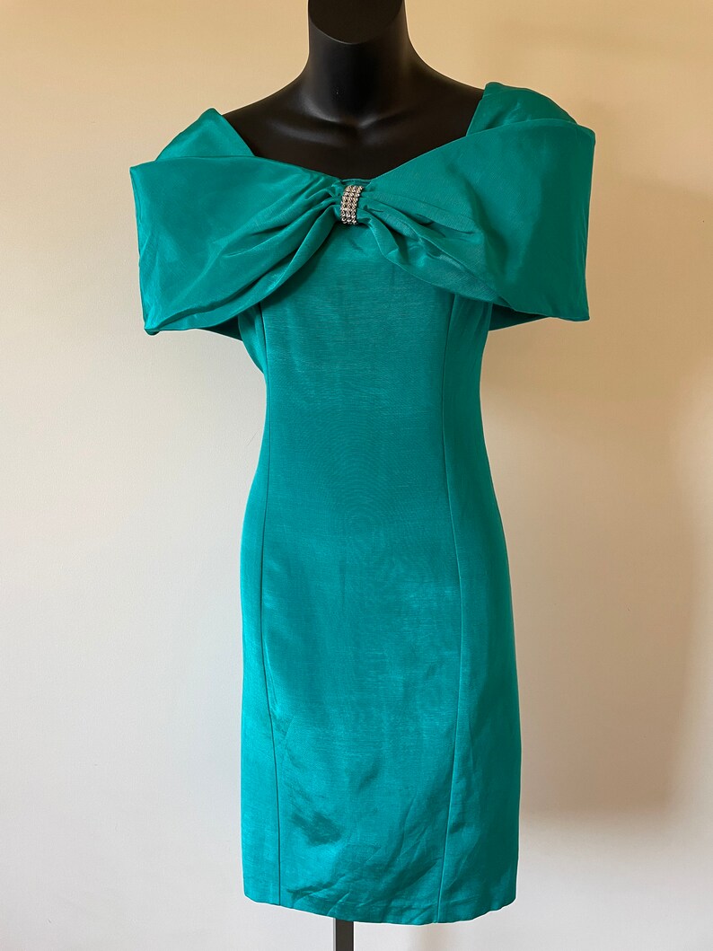 Puede incluir: Vestido de c&oacute;ctel color turquesa con escote de hombros descubiertos y un gran lazo en el busto. Un broche de pedrer&iacute;a brillante adorna el lazo. El vestido entallado, a la altura de la rodilla, se exhibe en un maniqu&iacute; negro.