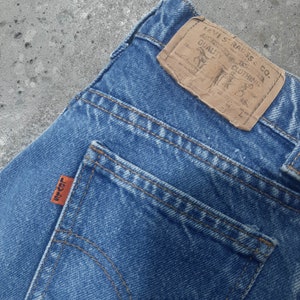 Puede incluir: Primer plano de unos vaqueros de mezclilla azules con una etiqueta de cuero marr&oacute;n y una etiqueta naranja. La etiqueta de cuero dice "LEVI STRAUSS & CO. QUALITY CLOTHING". La etiqueta naranja del bolsillo trasero dice "LEVI'S".
