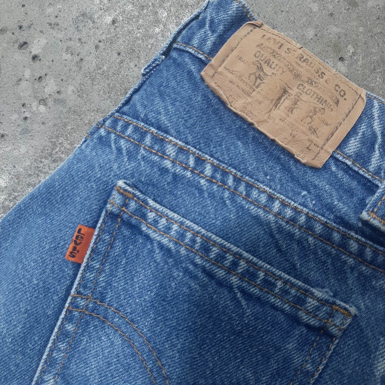 Puede incluir: Primer plano de unos vaqueros de mezclilla azules con una etiqueta de cuero marr&oacute;n y una etiqueta naranja. La etiqueta de cuero dice "LEVI STRAUSS & CO. QUALITY CLOTHING". La etiqueta naranja del bolsillo trasero dice "LEVI'S".