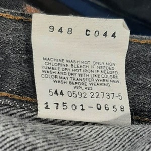 May include: Close-up of a white tag with black text attached to denim fabric. The tag includes care instructions and several numerical codes. The fabric is a dark wash denim.