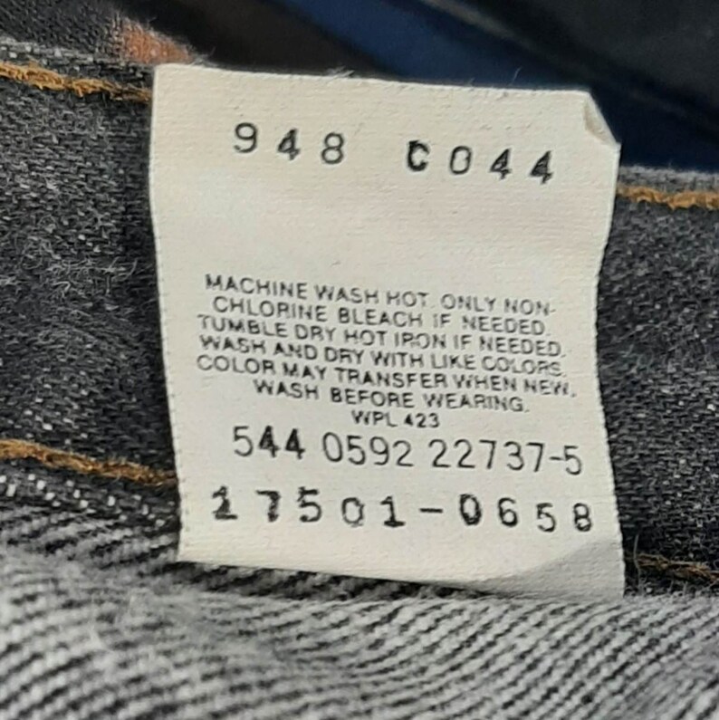 May include: Close-up of a white tag with black text attached to denim fabric. The tag includes care instructions and several numerical codes. The fabric is a dark wash denim.