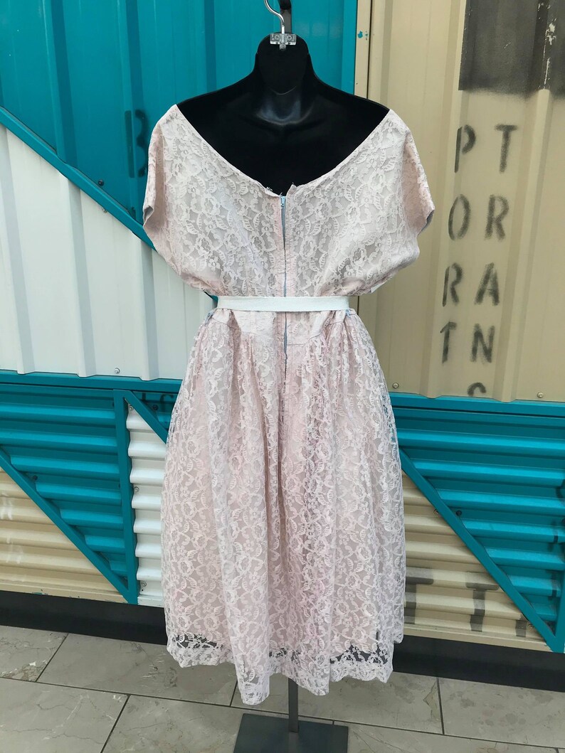 Puede incluir: Vestido de encaje rosa claro sobre un maniqu&iacute; negro. El vestido presenta un escote en V, mangas cortas y una falda a la rodilla. Un cintur&oacute;n blanco ci&ntilde;e la cintura. El vestido est&aacute; hecho de delicado tejido de encaje.