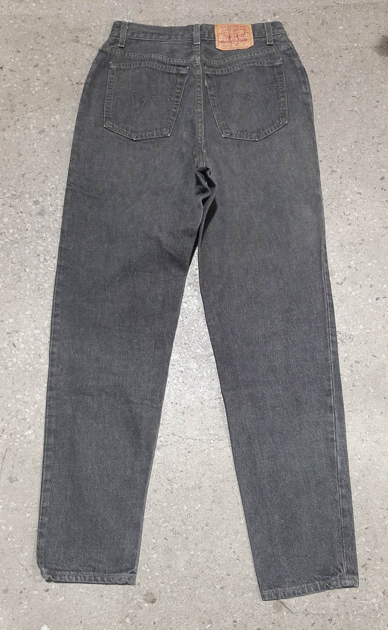 May include: Back view of a pair of charcoal gray denim jeans. The jeans have two back pockets and a small leather patch with red lettering. The jeans are a classic style, suitable for everyday wear.
