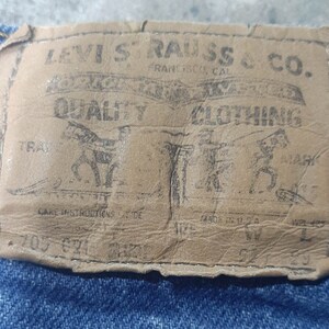 Puede incluir: Primer plano de una etiqueta de cuero desgastada de Levi Strauss & Co. cosida sobre mezclilla azul. La etiqueta presenta las palabras "QUALITY CLOTHING" y un gr&aacute;fico de dos caballos. La etiqueta tambi&eacute;n incluye el texto "FRANCISCO CAL" y "MADE IN U.S.A."