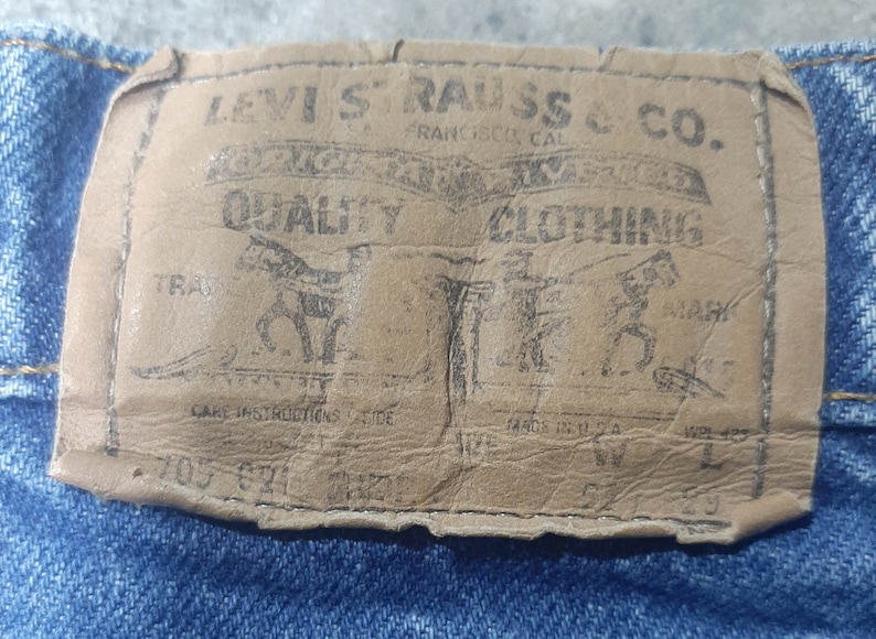 Puede incluir: Primer plano de una etiqueta de cuero desgastada de Levi Strauss & Co. cosida sobre mezclilla azul. La etiqueta presenta las palabras "QUALITY CLOTHING" y un gr&aacute;fico de dos caballos. La etiqueta tambi&eacute;n incluye el texto "FRANCISCO CAL" y "MADE IN U.S.A."