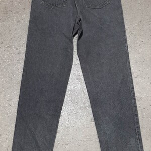 May include: A pair of dark gray denim jeans, viewed from the back. The jeans have two back pockets, a small red tag, and a leather patch. The jeans are straight-legged and appear to be vintage.