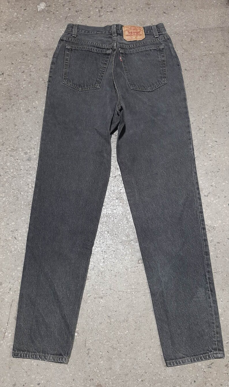 May include: A pair of dark gray denim jeans, viewed from the back. The jeans have two back pockets, a small red tag, and a leather patch. The jeans are straight-legged and appear to be vintage.