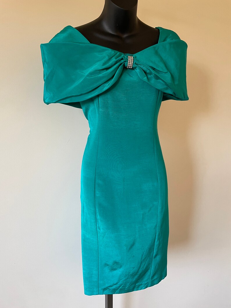 Puede incluir: Vestido turquesa con hombros descubiertos y un gran lazo en el escote, adornado con pedrer&iacute;a. El vestido tiene una silueta ajustada y se exhibe en un maniqu&iacute; negro.