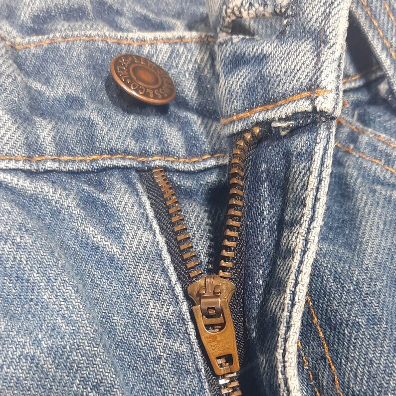 Puede incluir: Primer plano de unos vaqueros azules. La imagen destaca el bot&oacute;n, la cremallera y las costuras. El bot&oacute;n es de color cobre y la cremallera est&aacute; desabrochada, revelando los dientes y el tirador.