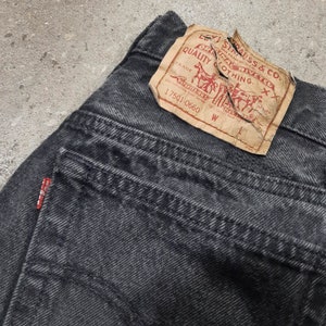 May include: Close-up of black denim jeans with a faded appearance. The jeans feature a red Levi's tag and a brown paper label with the Levi Strauss & Co. logo and text. The jeans are a classic style.