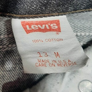 May include: Close-up of a Levi's tag, featuring the brand's logo in red. The tag reads "100% COTTON" and "13 M", with "MADE IN U.S.A." and "CARE ON REVERSE" printed below. The tag is attached to denim.