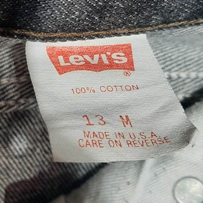 May include: Close-up of a Levi's tag, featuring the brand's logo in red. The tag reads "100% COTTON" and "13 M", with "MADE IN U.S.A." and "CARE ON REVERSE" printed below. The tag is attached to denim.