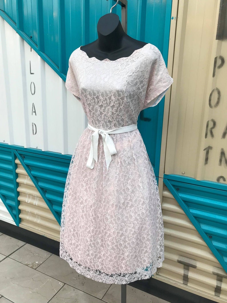 Puede incluir: Un vestido vintage de encaje rosa sobre un maniqu&iacute;. El vestido presenta un escote festoneado, mangas cortas y un cintur&oacute;n de cinta blanca. La falda hasta la rodilla est&aacute; completamente cubierta de encaje. El vestido se exhibe sobre un fondo de paneles turquesa y blancos.