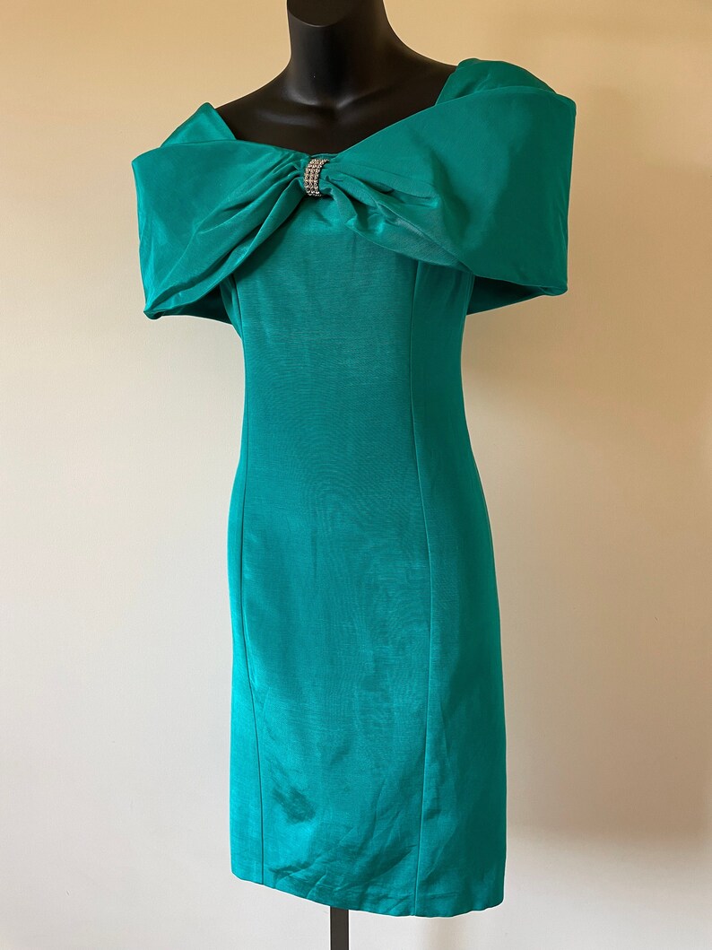 Puede incluir: Vestido turquesa con hombros descubiertos y un gran lazo en el escote, adornado con pedrer&iacute;a. El vestido tiene una silueta ajustada y un largo hasta la rodilla, ideal para eventos formales. El vestido mide unos 100 cm de largo.
