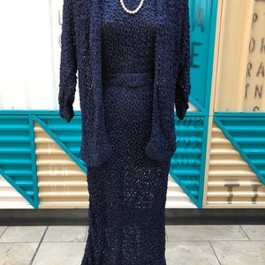 K&ouml;nnte beinhalten: Ein marineblaues, strukturiertes Strickkleid mit passender Jacke und G&uuml;rtel. Eine wei&szlig;e Perlenkette liegt um den Hals der Schaufensterpuppe. Das Kleid ist knielang und die Jacke hat Dreiviertel&auml;rmel. Das Outfit wird auf einem schwarzen Mannequin-St&auml;nder pr&auml;sentiert.