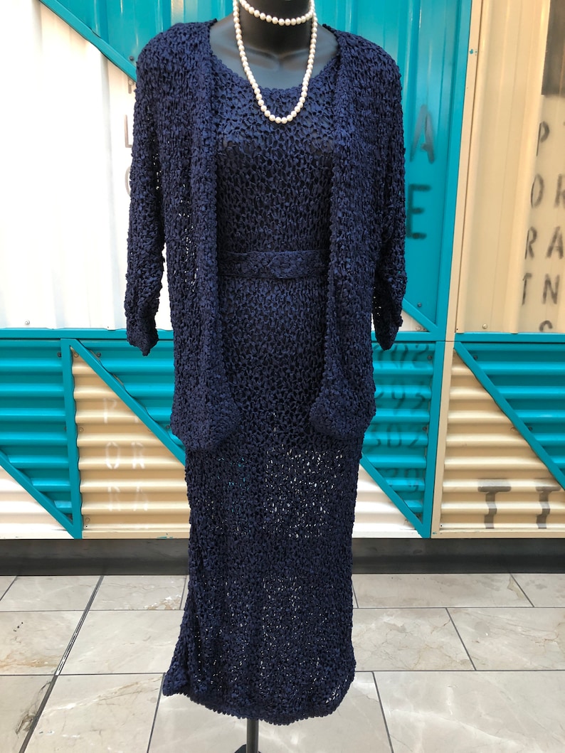 K&ouml;nnte beinhalten: Ein marineblaues, strukturiertes Strickkleid mit passender Jacke und G&uuml;rtel. Eine wei&szlig;e Perlenkette liegt um den Hals der Schaufensterpuppe. Das Kleid ist knielang und die Jacke hat Dreiviertel&auml;rmel. Das Outfit wird auf einem schwarzen Mannequin-St&auml;nder pr&auml;sentiert.