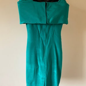 Puede incluir: Un vestido turquesa con hombros descubiertos. El vestido presenta un escote estructurado y plegado, y una silueta ajustada. La tela tiene un brillo sutil y una abertura visible en el dobladillo trasero. El vestido se exhibe en un maniqu&iacute; negro.