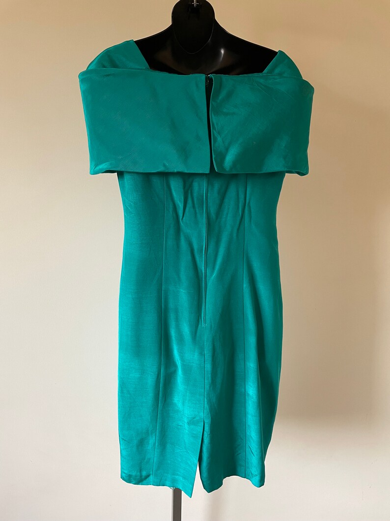 Puede incluir: Un vestido turquesa con hombros descubiertos. El vestido presenta un escote estructurado y plegado, y una silueta ajustada. La tela tiene un brillo sutil y una abertura visible en el dobladillo trasero. El vestido se exhibe en un maniqu&iacute; negro.