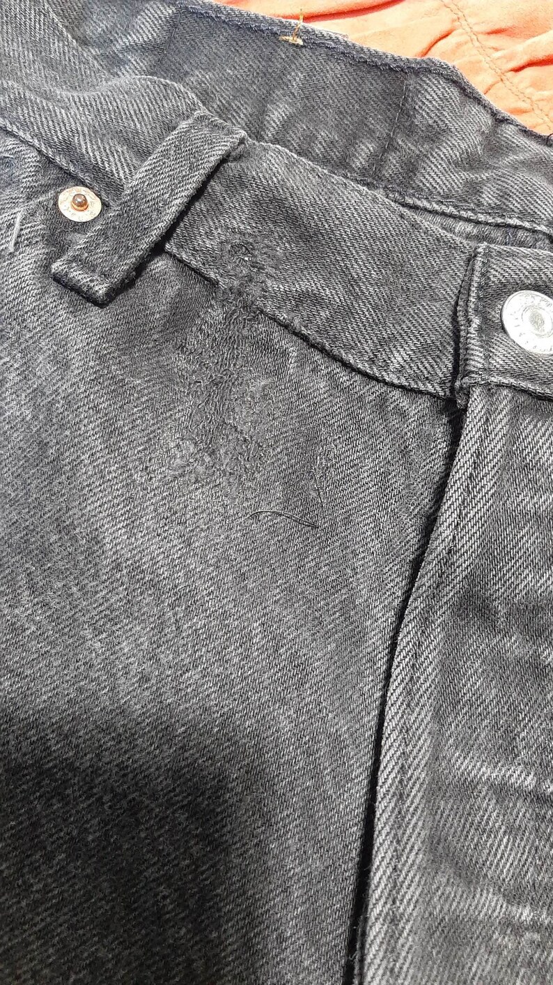 May include: Close-up of black denim jeans, showing the waistband, belt loop, and button closure. The fabric has a textured appearance with visible stitching and a worn look. The jeans are a classic style.
