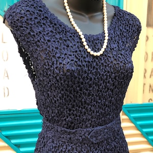 K&ouml;nnte beinhalten: Ein marineblaues Vintage-Kleid mit einem strukturierten, offen gewebten Design. Das Kleid hat eine taillierte Taille und kurze &Auml;rmel. Eine Kette aus wei&szlig;en Perlen ist um den Hals der Schaufensterpuppe drapiert.