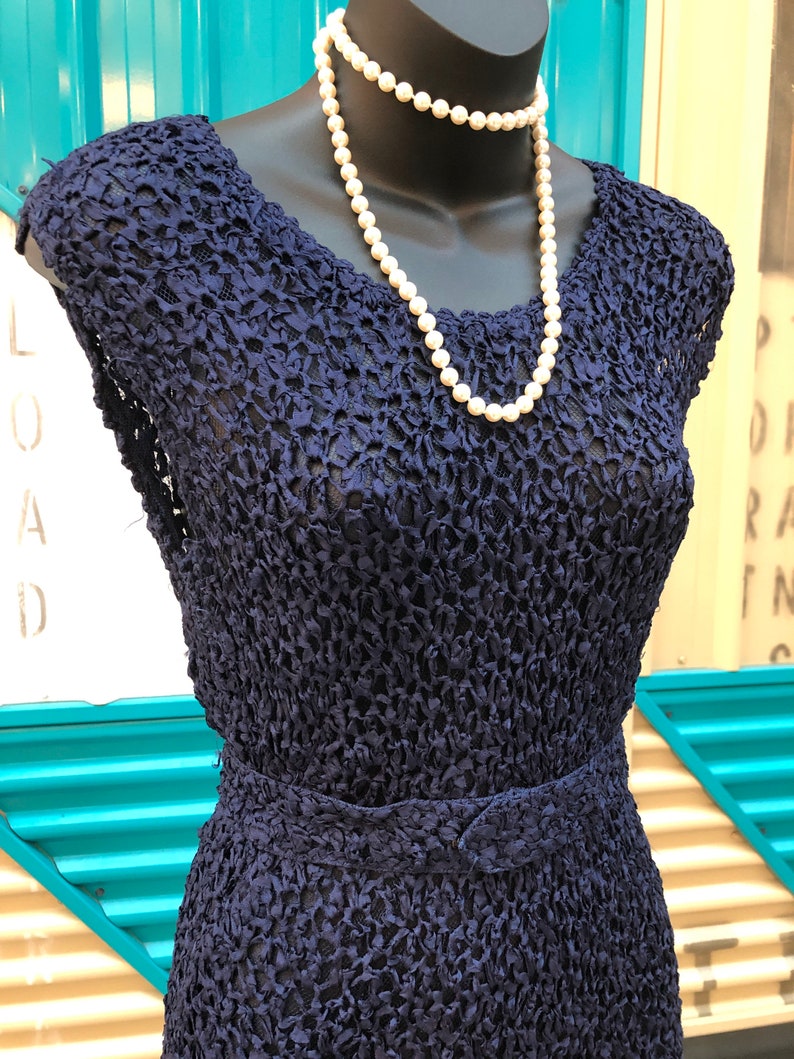 K&ouml;nnte beinhalten: Ein marineblaues Vintage-Kleid mit einem strukturierten, offen gewebten Design. Das Kleid hat eine taillierte Taille und kurze &Auml;rmel. Eine Kette aus wei&szlig;en Perlen ist um den Hals der Schaufensterpuppe drapiert.