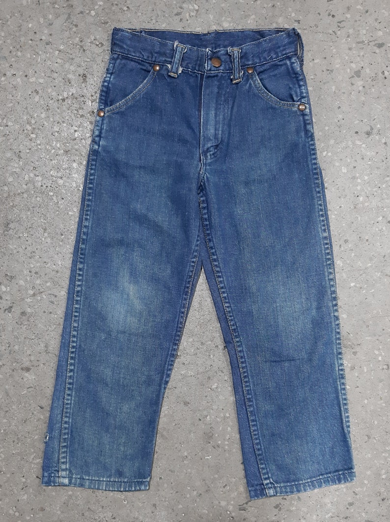 May include: A pair of blue denim jeans with a classic five-pocket design. The jeans have a button and zipper closure, belt loops, and copper rivets. The denim has a faded appearance, suggesting wear. The jeans are likely for children.