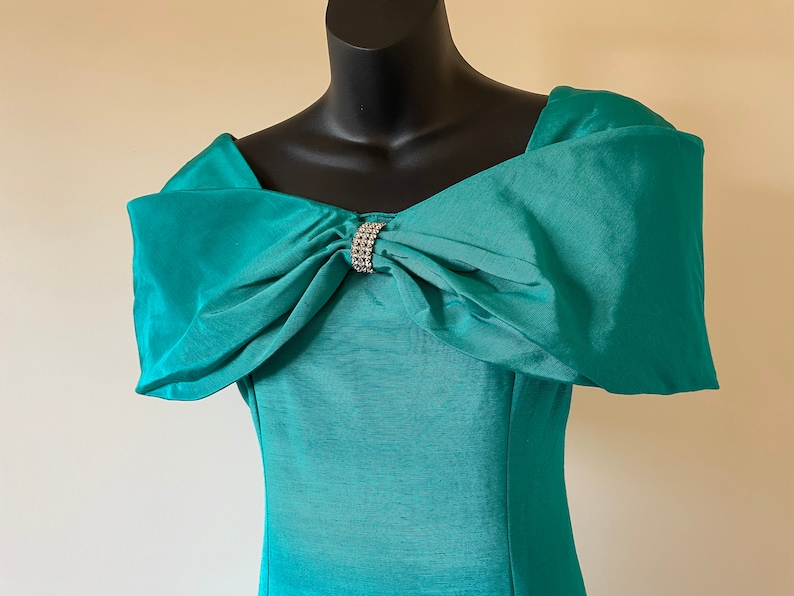 Puede incluir: Vestido turquesa con hombros descubiertos y un gran lazo en el escote, adornado con pedrer&iacute;a. El vestido se exhibe en un maniqu&iacute; negro. El color es un turquesa vibrante.