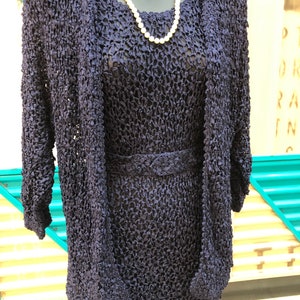 K&ouml;nnte beinhalten: Ein marineblaues, strukturiertes Strickkleid mit passender Jacke und G&uuml;rtel. Eine wei&szlig;e Perlenkette ist um den Hals der Schaufensterpuppe drapiert. Das Kleid und die Jacke haben ein einzigartiges, offenes Webdesign.