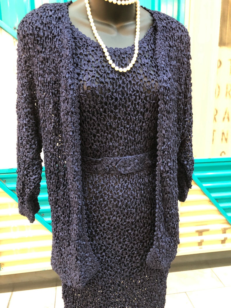 K&ouml;nnte beinhalten: Ein marineblaues, strukturiertes Strickkleid mit passender Jacke und G&uuml;rtel. Eine wei&szlig;e Perlenkette ist um den Hals der Schaufensterpuppe drapiert. Das Kleid und die Jacke haben ein einzigartiges, offenes Webdesign.