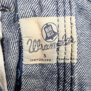 May include: Close-up of a Wrangler jeans label. The label is white with blue text and a bell logo. The text reads "Wrangler", "5", "SANFORIZED", and "MADE IN U.S.A.". The jeans are a light blue denim.