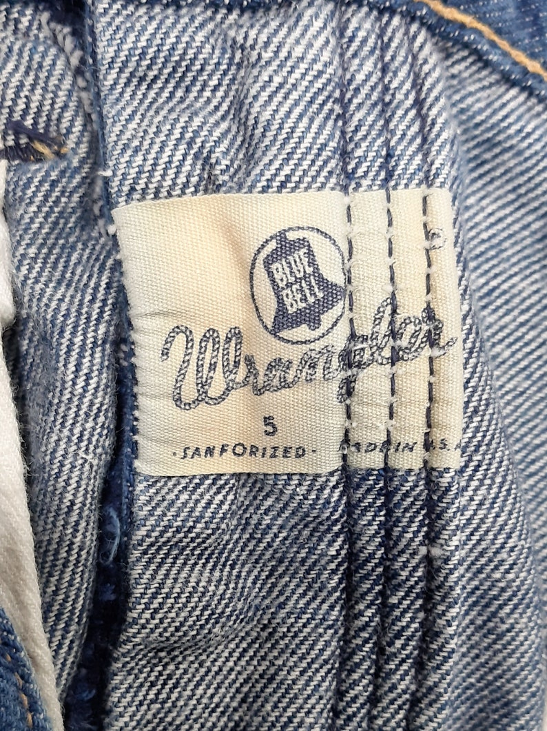 May include: Close-up of a Wrangler jeans label. The label is white with blue text and a bell logo. The text reads "Wrangler", "5", "SANFORIZED", and "MADE IN U.S.A.". The jeans are a light blue denim.