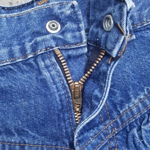 Puede incluir: Primer plano de un pantal&oacute;n corto vaquero azul. El pantal&oacute;n corto tiene una cremallera met&aacute;lica, un cierre de bot&oacute;n y costuras visibles en hilo naranja-marr&oacute;n contrastante. La tela vaquera est&aacute; ligeramente descolorida.