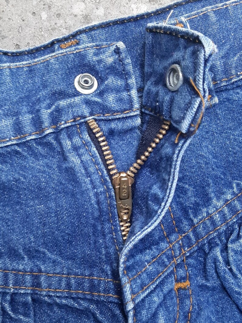 Puede incluir: Primer plano de un pantal&oacute;n corto vaquero azul. El pantal&oacute;n corto tiene una cremallera met&aacute;lica, un cierre de bot&oacute;n y costuras visibles en hilo naranja-marr&oacute;n contrastante. La tela vaquera est&aacute; ligeramente descolorida.