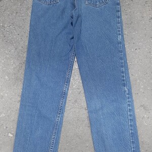 Puede incluir: Vista trasera de unos vaqueros de mezclilla azules con un dise&ntilde;o cl&aacute;sico de cinco bolsillos. Incluye un parche de cuero marr&oacute;n con letras en relieve y costuras naranjas en los bolsillos traseros. Los vaqueros est&aacute;n planos, mostrando el corte de pierna recta.