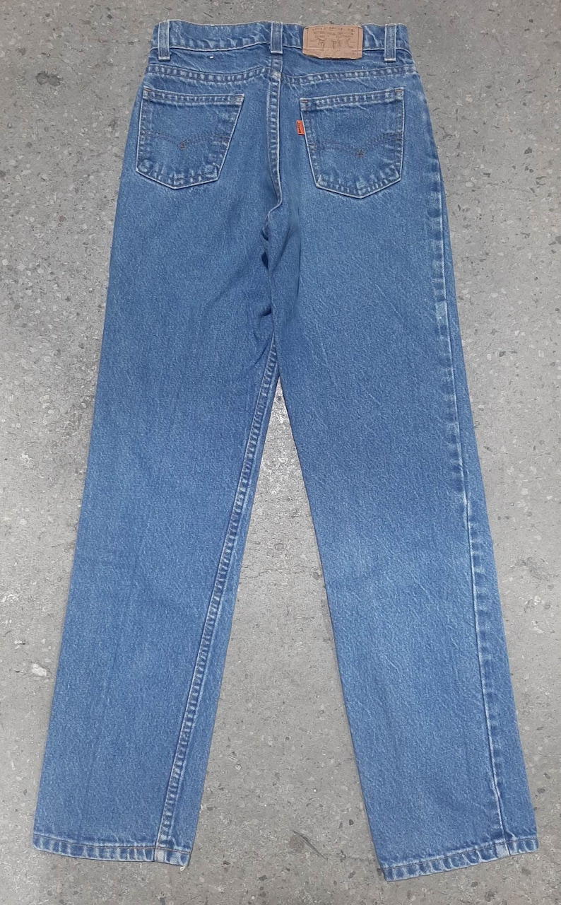 Puede incluir: Vista trasera de unos vaqueros de mezclilla azules con un dise&ntilde;o cl&aacute;sico de cinco bolsillos. Incluye un parche de cuero marr&oacute;n con letras en relieve y costuras naranjas en los bolsillos traseros. Los vaqueros est&aacute;n planos, mostrando el corte de pierna recta.