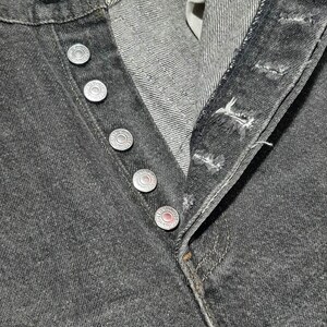 May include: Close-up of black denim jeans with a button fly. The jeans have silver-colored metal buttons and rivets. The fabric shows a textured weave. The jeans are unbuttoned, revealing the interior construction.