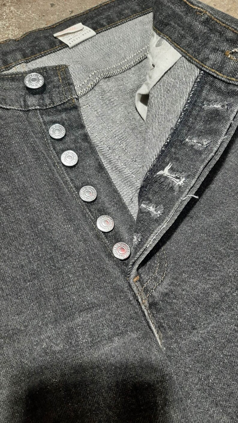May include: Close-up of black denim jeans with a button fly. The jeans have silver-colored metal buttons and rivets. The fabric shows a textured weave. The jeans are unbuttoned, revealing the interior construction.