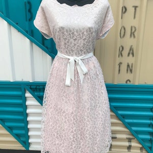 Puede incluir: Vestido de encaje rosa claro con escote festoneado y mangas cortas. El vestido presenta un cintur&oacute;n de cinta blanca y una falda a la rodilla. El vestido se exhibe en un maniqu&iacute; negro sobre un fondo de paneles turquesa y beige.