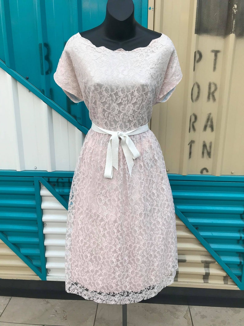 Puede incluir: Vestido de encaje rosa claro con escote festoneado y mangas cortas. El vestido presenta un cintur&oacute;n de cinta blanca y una falda a la rodilla. El vestido se exhibe en un maniqu&iacute; negro sobre un fondo de paneles turquesa y beige.