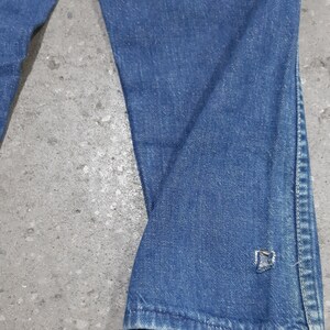 May include: Close-up of blue denim jeans. The jeans have a slight flare at the bottom and a small tear near the hem. The fabric shows signs of wear and tear, with visible fading and creases.
