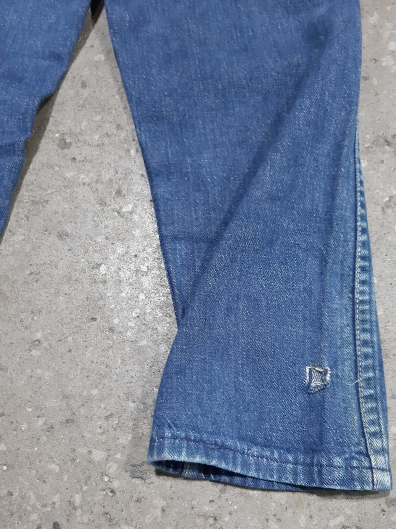 May include: Close-up of blue denim jeans. The jeans have a slight flare at the bottom and a small tear near the hem. The fabric shows signs of wear and tear, with visible fading and creases.