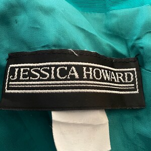 Puede incluir: Primer plano de una prenda de color turquesa con una etiqueta negra. La etiqueta presenta el texto bordado "JESSICA HOWARD" en blanco. La prenda tiene una cremallera y una tela texturizada.