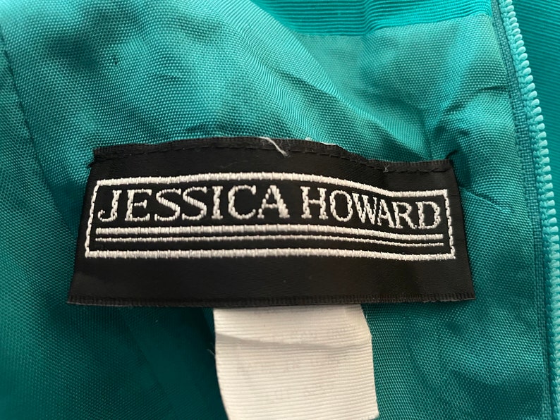Puede incluir: Primer plano de una prenda de color turquesa con una etiqueta negra. La etiqueta presenta el texto bordado "JESSICA HOWARD" en blanco. La prenda tiene una cremallera y una tela texturizada.