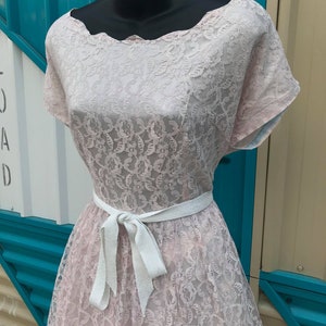 Puede incluir: Vestido vintage de encaje rosa con escote festoneado y mangas cortas. El vestido presenta un cintur&oacute;n de cinta blanca atado con un lazo en la cintura. El vestido se exhibe en un maniqu&iacute; negro.