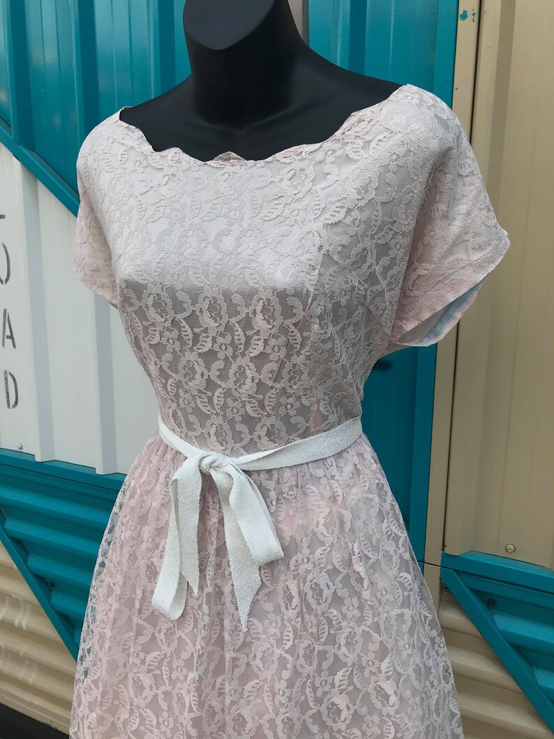 Puede incluir: Vestido vintage de encaje rosa con escote festoneado y mangas cortas. El vestido presenta un cintur&oacute;n de cinta blanca atado con un lazo en la cintura. El vestido se exhibe en un maniqu&iacute; negro.