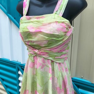 Peut inclure: Robe florale vintage, vert clair et rose, présentée sur un mannequin noir. La robe présente un corsage froncé, un détail de taille nouée et de larges bretelles. Le tissu est délicat et semi-transparent, typique des robes vintage.
