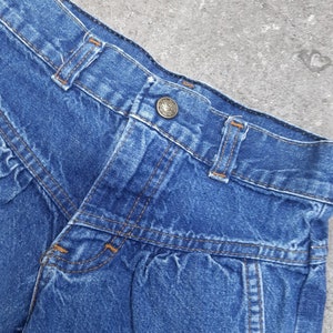 Puede incluir: Primer plano de unos pantalones cortos vaqueros azules. Los pantalones cortos tienen cierre de bot&oacute;n, trabillas y costuras decorativas. El denim tiene un aspecto ligeramente descolorido, lo que sugiere un estilo vintage. Los pantalones cortos son una prenda cl&aacute;sica e informal.